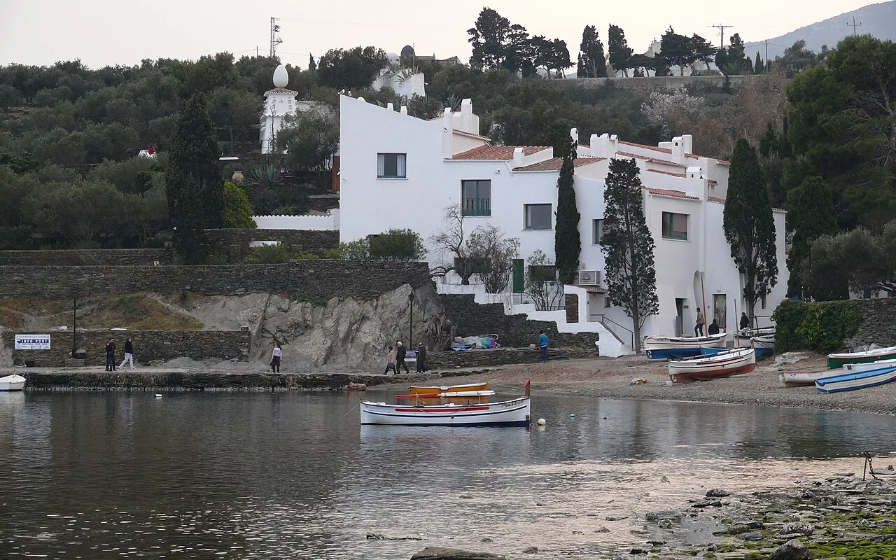 Dalí's whitewashed house at Portlligat seen from the beach, cypresses beside its stairs and fishing boats on the shore